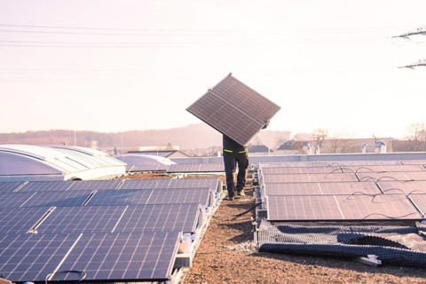 A worker carries a solar panel over a roof during sunset, PV assembly at OBI Baumarkt, Mühlacker,