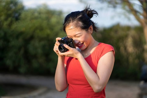 Asian woman in a red shirt capturing moments outdoors with a digital camera. Her joyful expression