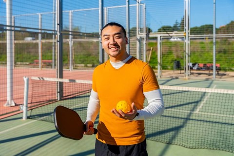 Asian man smiling on outdoor pickleball court, holding paddle and perforated yellow ball, ready to
