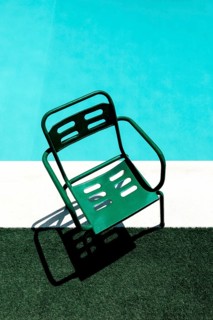 Top view of a green metal chair casting a shadow on artificial grass beside a pool. The vibrant