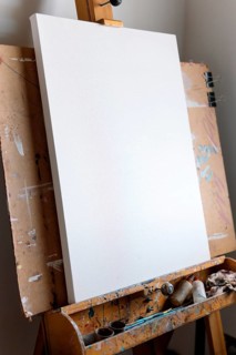 A clean, blank canvas rests on a well-used wooden easel covered in paint splatters. Art supplies