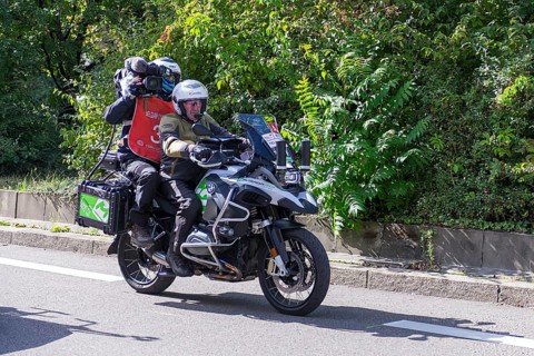 A cameraman on a motorbike rides during a bicycle race on a sunny road surrounded by green nature,
