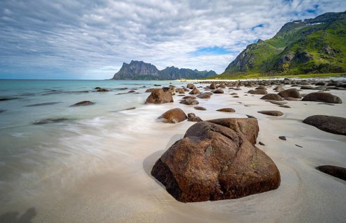 Rocks on Uttakleiv sandy beach, coast and blue sea, long exposure, rocky mountain peaks,