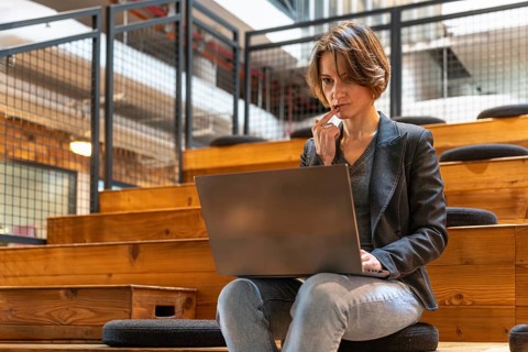 Focused young woman in jeans and blazer thinking deeply while working on laptop in modern coworking