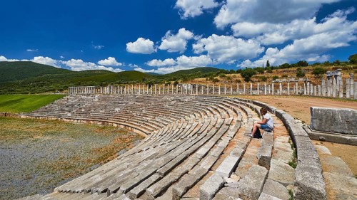 Person sitting in ancient ruins of a stadium under blue sky with clouds, Messene Stadium, Ancient