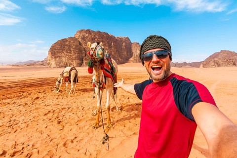 Smiling man traveler taking selfie standing near saddled camel orange sand valley sandstone rock