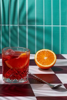 A drink with ice and orange slices is placed in a glass on a checkered table. A cut orange sits
