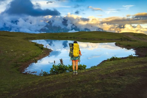 Woman hiker with yellow backpack stand by mirror-like Koruldi Lake enjoy sunset in Georgia. A