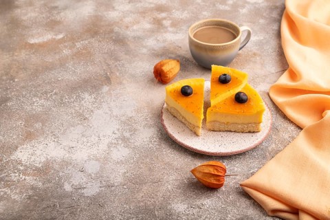 Cheesecake with mango cream, cup of coffee on brown concrete background with orange textile, copy