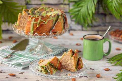 A sliced cake and pieces of cake with green icing next to a cup of coffee and almonds