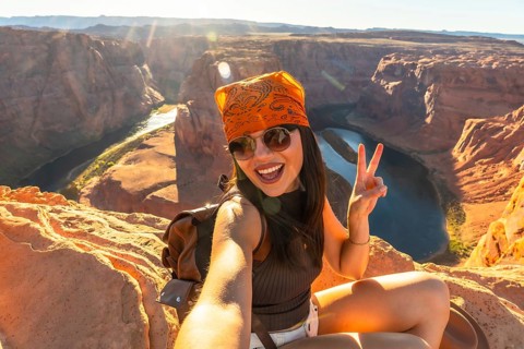 Young woman traveler at horseshoe bend in arizona taking a joyful selfie with a peace sign, sitting