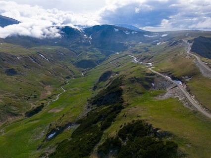 Road runs through green mountain hills in a vast valley under cloudy sky, aerial view, near Rânca,