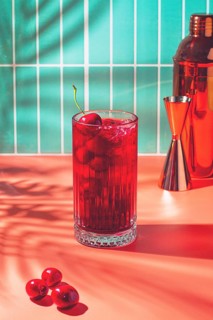 A bright drink made with cherry sits on a table next to some fresh cherries. The background shows a