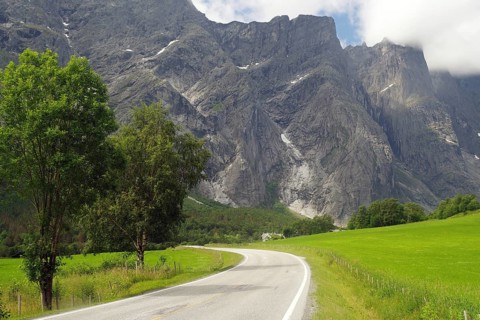 Winding country road between green fields and dramatic mountains under cloudy sky, Tom Cruise