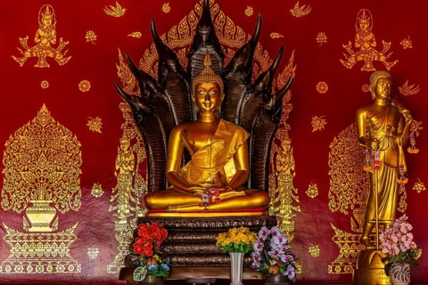 Gilded seated Buddha statue in front of artfully decorated red temple wall, Buddhist temple complex