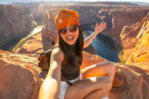 Young happy woman sitting on a cliff taking a selfie with horseshoe bend and the colorado river in