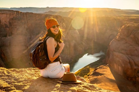 Young woman traveler with backpack and bandana smiling, sitting on a rocky cliff overlooking the