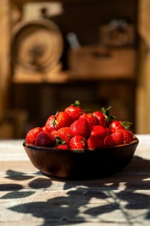 Bowl filled with ripe strawberries on wooden table under bright sunlight. Hard shadows fall around
