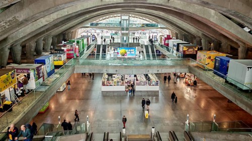 Panorama of large, modern station concourse with concrete walls and people on escalators, Oriente