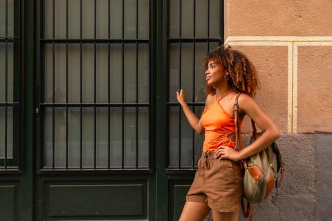 Young woman with afro hair, wearing an orange top and brown shorts, standing confidently in an