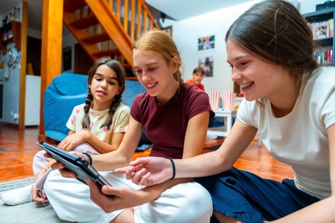 Three smiling children sit on the living room floor as two girls share a tablet, pointing at the