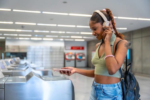Young woman smiling, wearing headphones and a backpack, using her smartphone to pay and pass