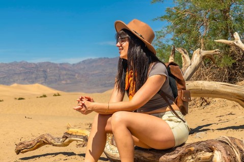 Woman wearing a hat and backpack sitting on dry wood and looking at sand dunes with mountains in