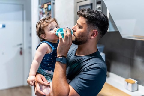 Father gently helps toddler son drink from a sippy cup in the kitchen, capturing a tender moment of