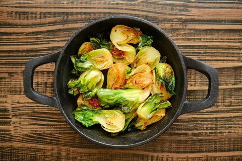 Bok choy is being cooked in a cast iron skillet. The greens are heated and show signs of roasting