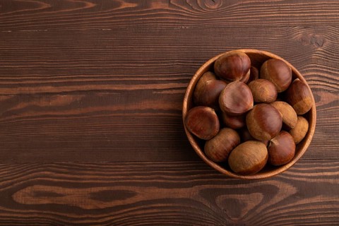 Wooden bowl with raw edible ?hestnuts on brown wooden background, top view, flat lay, copy space,