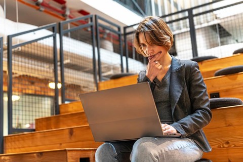 Thoughtful young woman in jeans and blazer resting hand on face while working on laptop in
