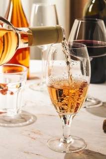 A person is pouring wine from a bottle into a clear glass. There are other drinks on the table,
