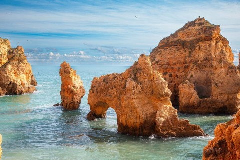 A fascinating rock arch juts out of the blue sea, surrounded by cliffs and clear skies, rocky