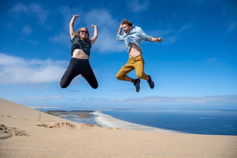 Tourists, young couple jumping in the air, stretching arms in the air, view of high dunes of the