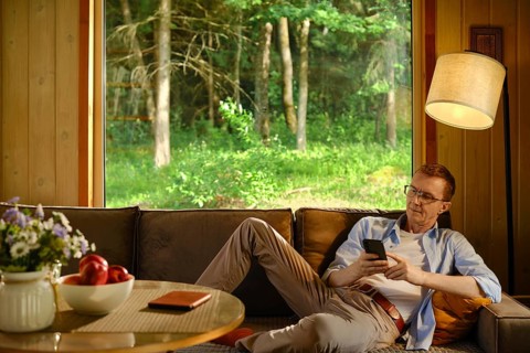 Middle-aged man rests on sofa inside wooden cabin with air conditioner and looks at smartphone