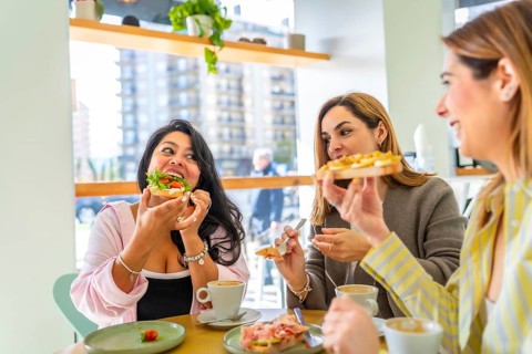 Three diverse female friends sitting at a table in a bright cafe, laughing and eating a fresh