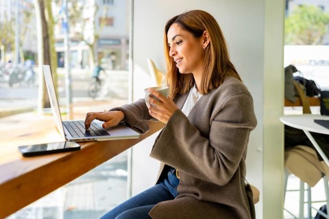 Young smiling woman sitting at a bar table in a cafe, typing on a laptop and holding a cup of