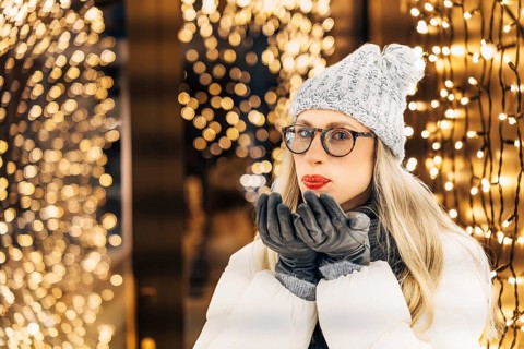 Young woman in knitted hat and down jacket blowing on gloved palms making a wish. Woman wearing