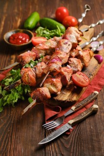 Shish kebob skewers arranged on a wooden board. Fresh tomatoes and cucumbers are placed around the