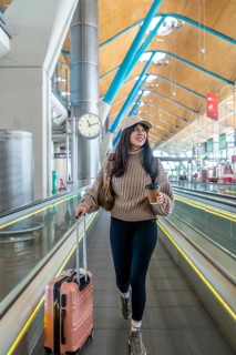 Young woman standing on an automatic moving walkway with a suitcase and a coffee cup, looking happy