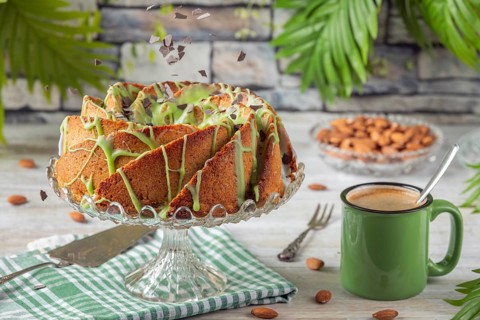 Chocolate chips fall on cake with green icing next to a cup of coffee and almonds