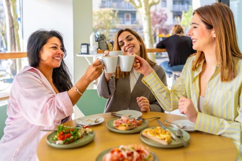 Three young women in a cafe raise coffee cups for a cheerful brunch toast, smiling and chatting