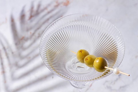 A clear glass cocktail sits on a white marble surface. Three green olives are placed on a skewer