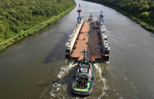 A tugboat pulls a barge on a tree-lined river, tugboat Bremerhaven pulls Dock V through the Kiel