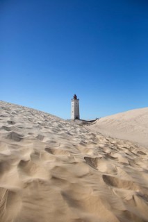 A lighthouse rises amidst wide sand dunes under a bright blue sky, lighthouse in the Rubjerg Knude