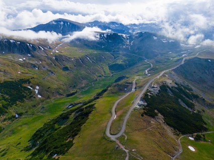 Serpentine road snakes through green mountain landscape under cloudy sky, aerial view, near Rânca,