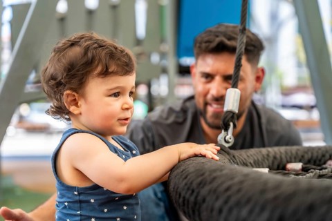 Dad smiling as toddler reaches for the swing at a sunny playground, sharing a playful, caring