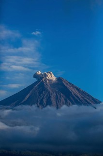 Smoke rises from the volcano as surrounding clouds and a blue sky frame the scene, Smoking Volcano,