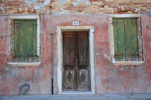 Two windows and a door in an old, weathered red brick façade, windows with green shutters, colorful