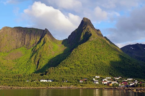 A village at the foot of a green mountain under a cloudy sky. Various houses are visible,